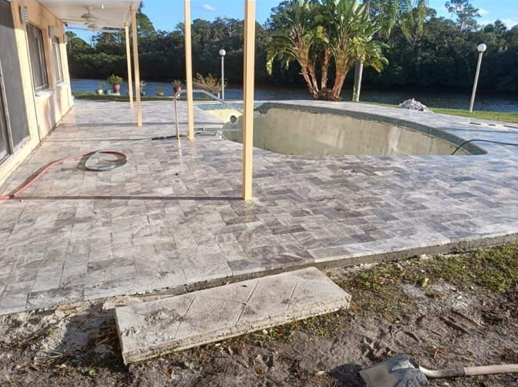 Patio with gray pavers, partially empty pool, construction materials, and a building on a sunny day.