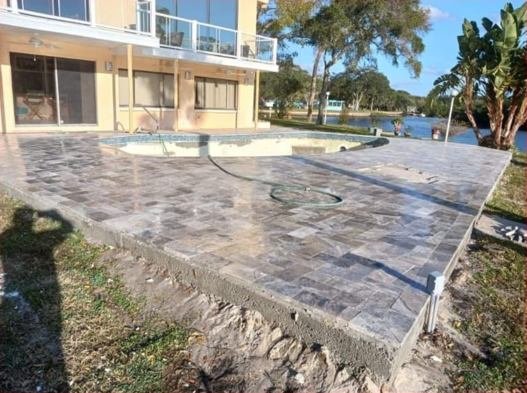 A new patio with pavers surrounds a pool. A home is in the background next to a body of water.