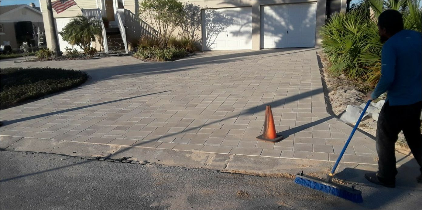 A person sweeping a brick driveway with a broom next to an orange traffic cone.