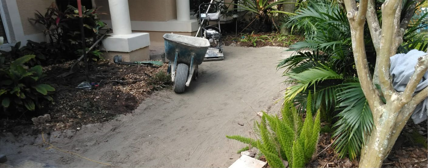 A wheelbarrow sits on a concrete path in a garden, near a house and trees.