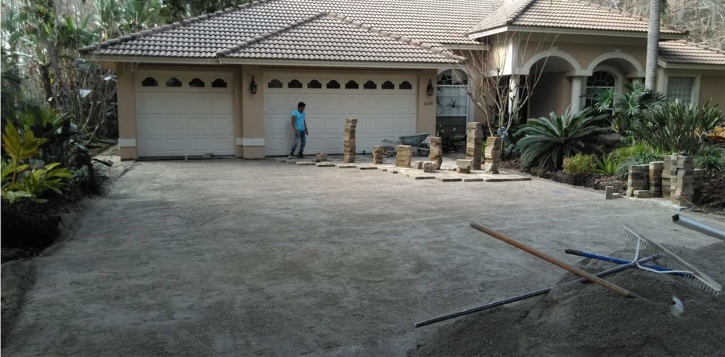 A person standing near a two-car garage, working on a gravel driveway in front of a house.