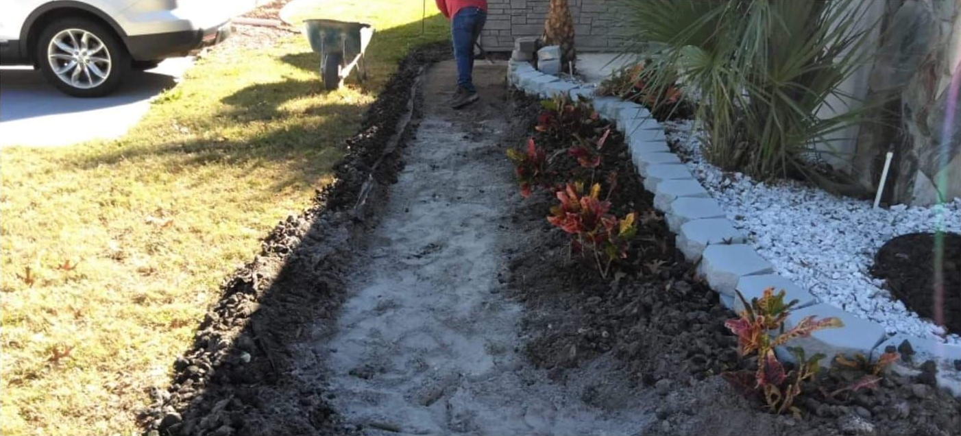 A narrow, excavated pathway bordered by grass and a garden bed with plants, white stones, and a wall.