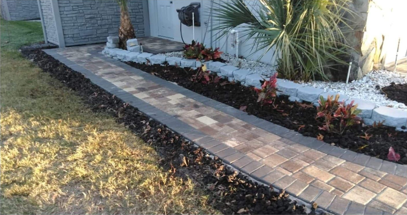 Brick walkway bordered by dark mulch and planting beds leads to a white building.