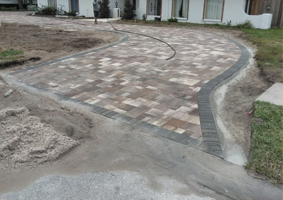 Driveway paved with multi-colored bricks, bordered by darker gray bricks; a house in the background.