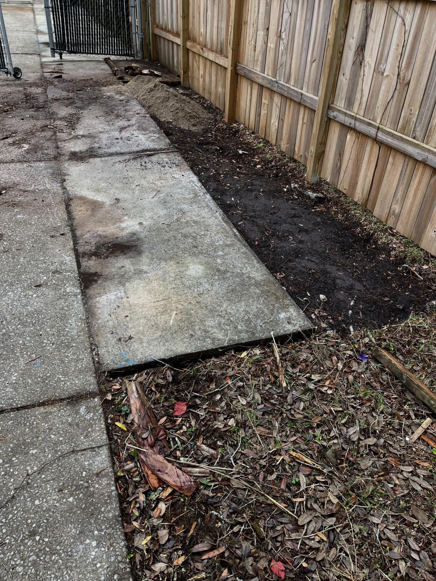 Concrete pathway next to a wooden fence and dirt garden bed.