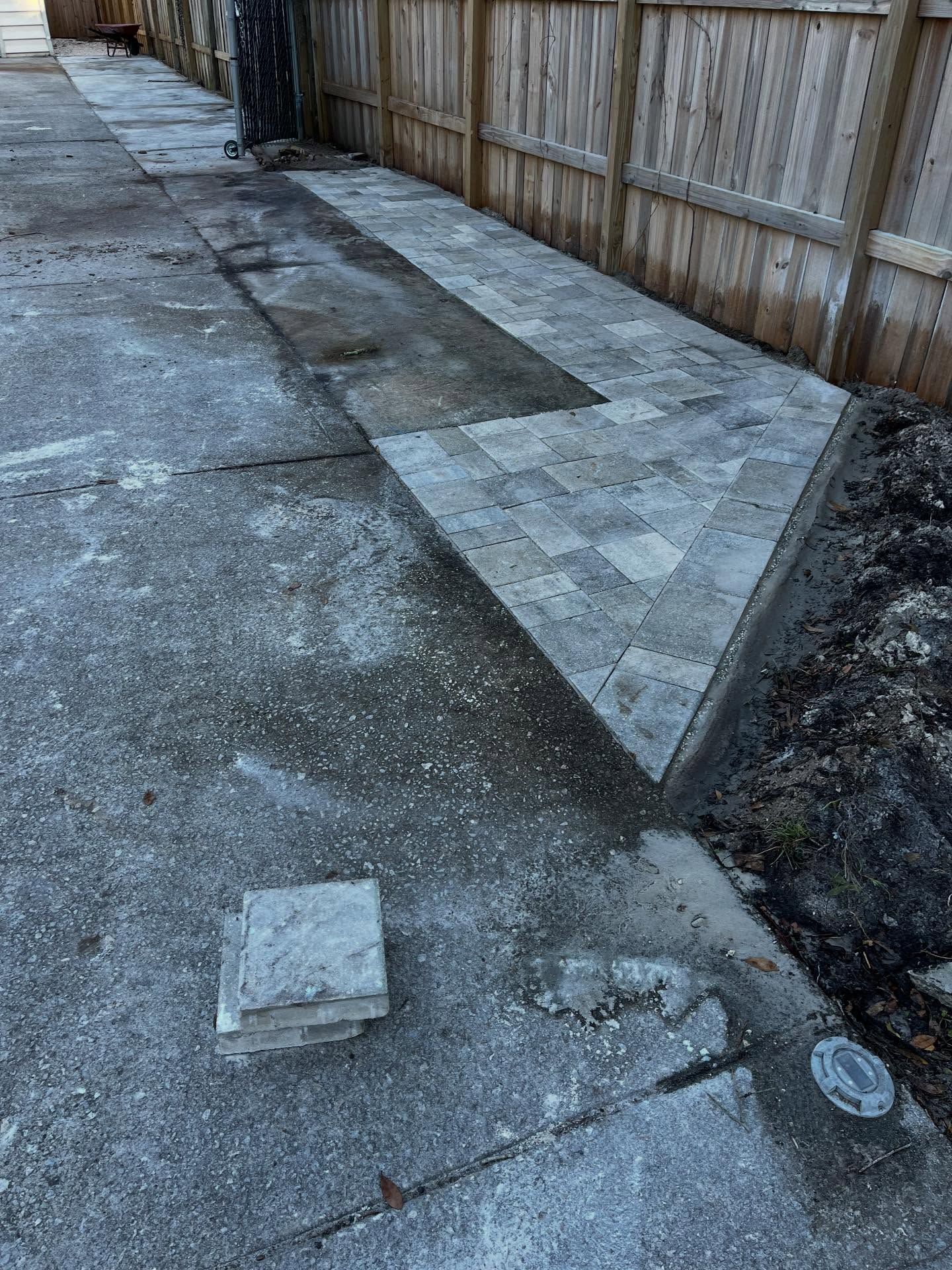 Concrete and newly paved walkway with pavers next to a wooden fence. Some pavers are stacked.
