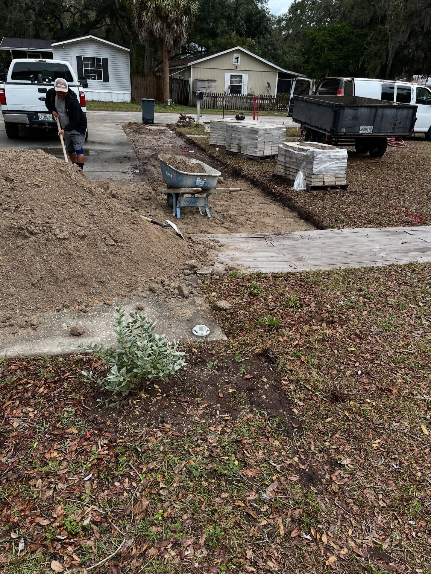 Construction site in a residential area. A person shovels dirt. Cement blocks and a wheelbarrow are present.