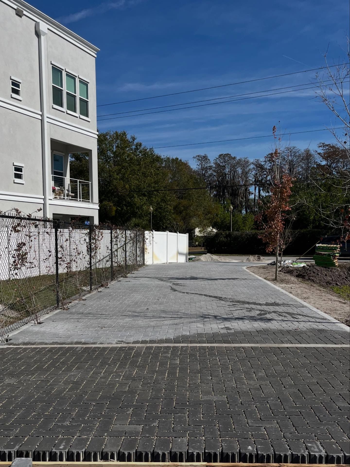 Gray brick parking area next to a white building and fence, with trees in the background under a blue sky.