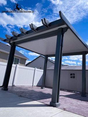 A pergola is sitting on top of a concrete patio next to a white fence.