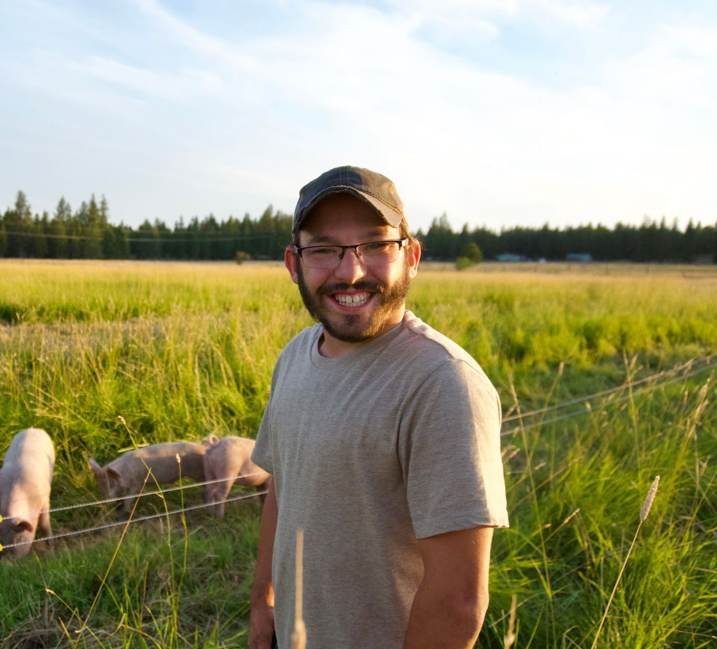 Man in a cap and glasses smiles in a field with pigs.