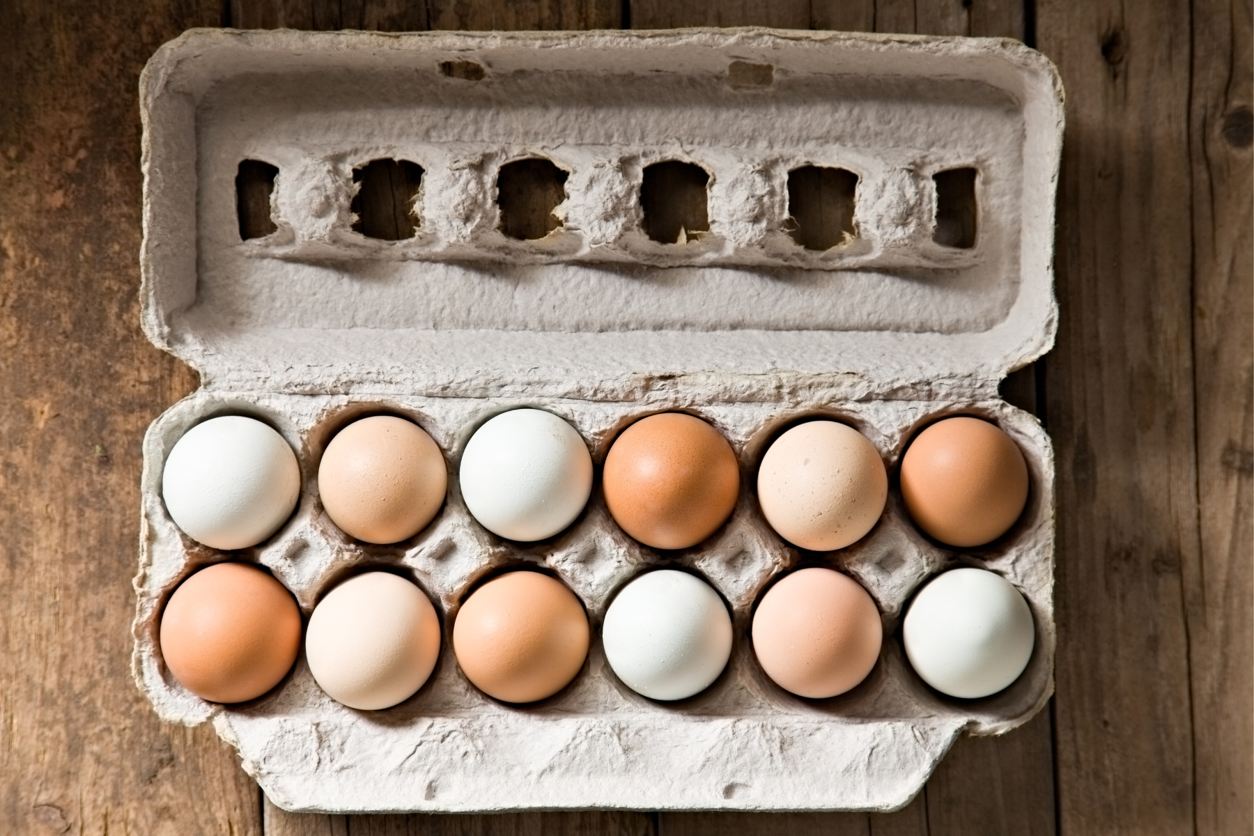 Cardboard egg carton with eleven eggs, some white and some brown, on a wood surface.