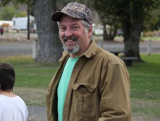 Man in camouflage cap, brown jacket, and teal shirt, smiling outdoors.