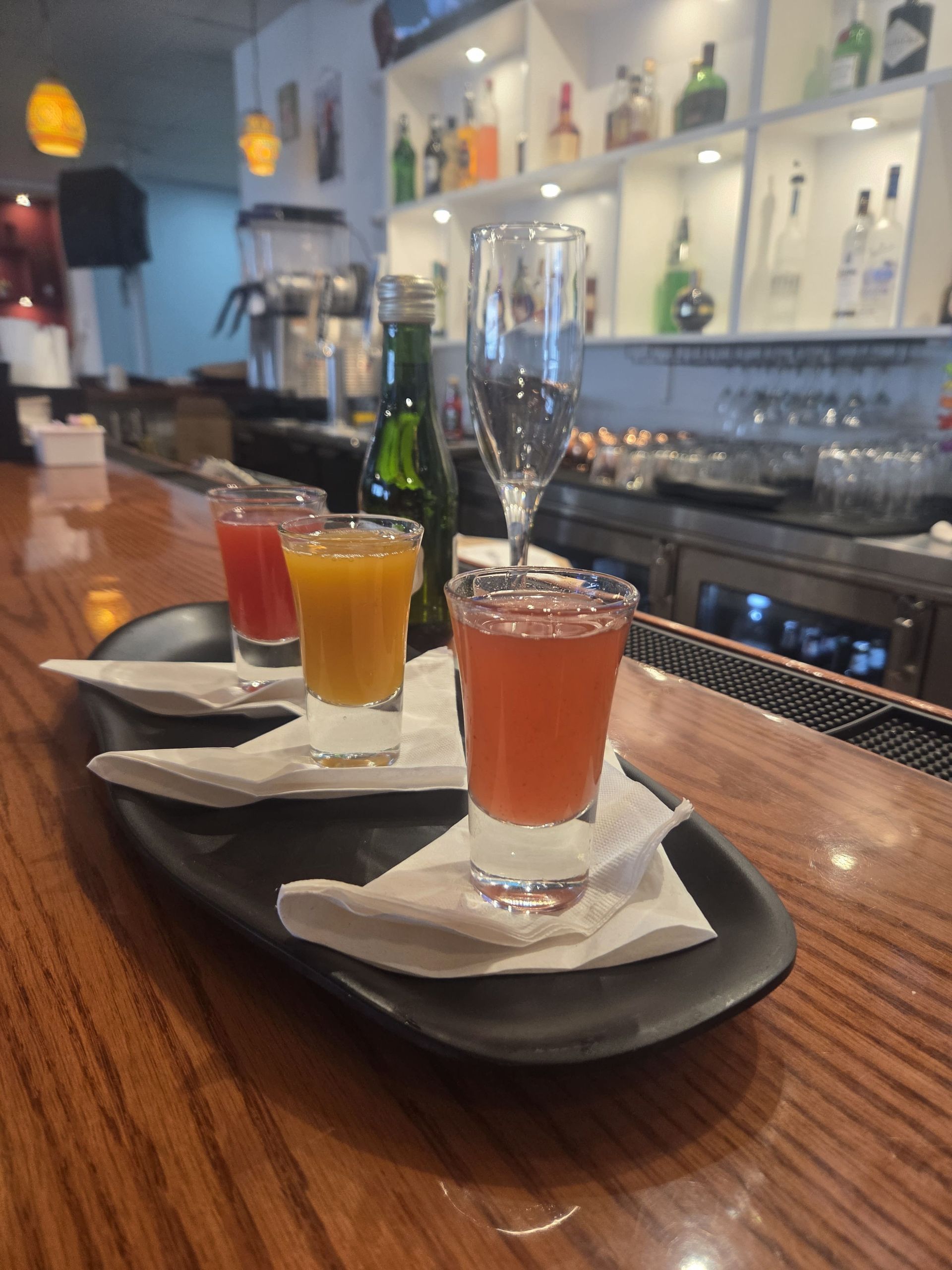 Shot glasses with colorful drinks on a black tray at a bar, bottle behind.