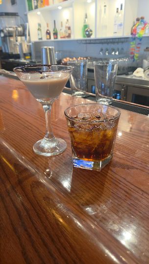 Two cocktails on a wooden bar: a martini with a garnish and a rocks glass with ice.