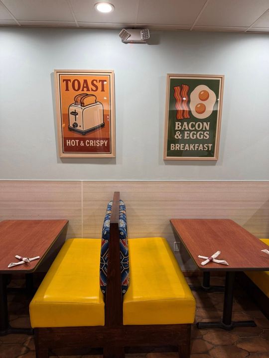 Restaurant booth with yellow seating, wood tables, and breakfast-themed pixel art posters.