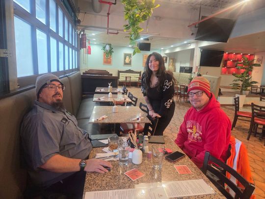 Three people at a restaurant table: two men seated, one woman standing behind them. Bright interior.
