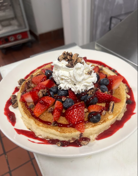 Pancake topped with strawberries, blueberries, whipped cream, and nuts, drizzled with red sauce on a white plate.