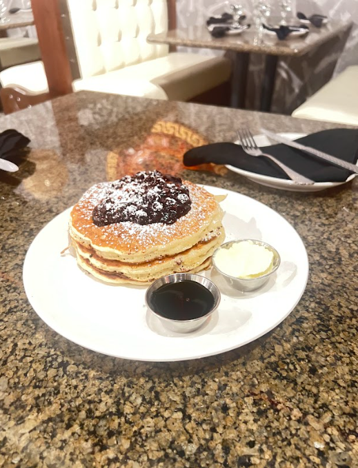 Stack of pancakes with blueberry compote, syrup, and butter on a white plate on a speckled table.