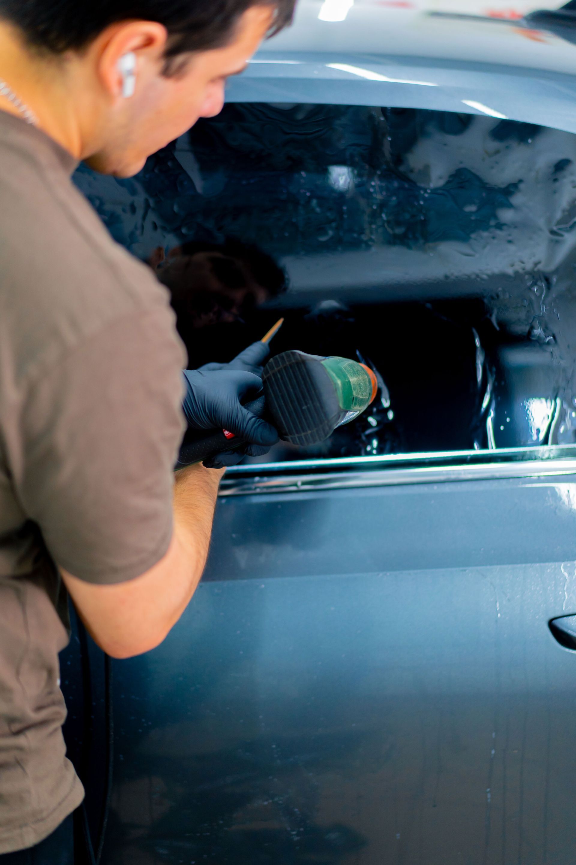 Person applying tint to a car window, using a tool and wearing gloves, in a garage.