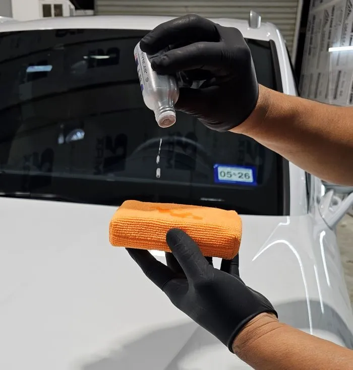 Hands in black gloves applying liquid from a bottle to an orange applicator sponge in front of a white car.