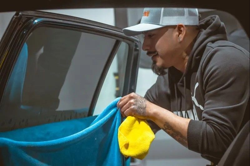 Man wiping car window with a yellow cloth. Wearing a hat and hoodie, indoors.