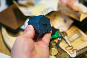 A person is holding a small model house in their hand in front of a pile of money.