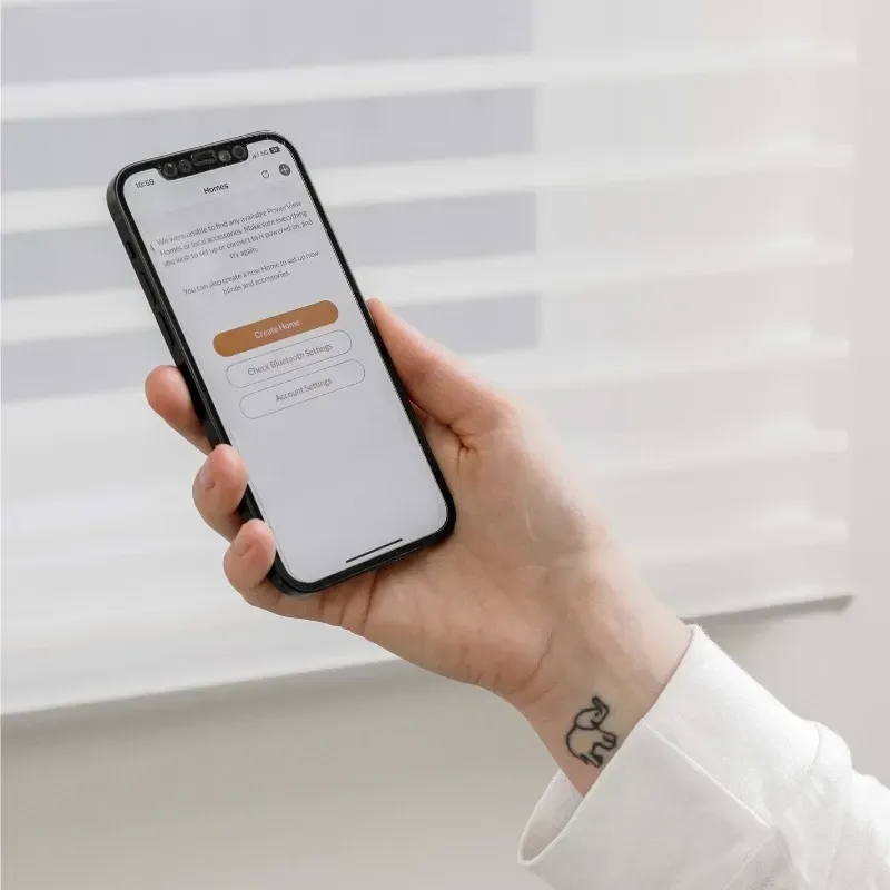 Hand holding a smartphone displaying a setup screen. Blinds are in the background.