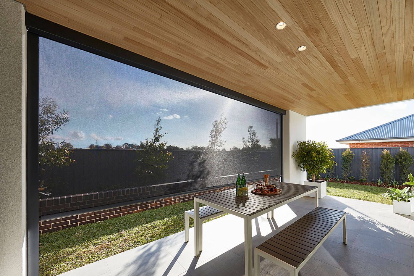 Outdoor patio with a dining table and benches, protected by a retractable screen, with trees and sky visible.