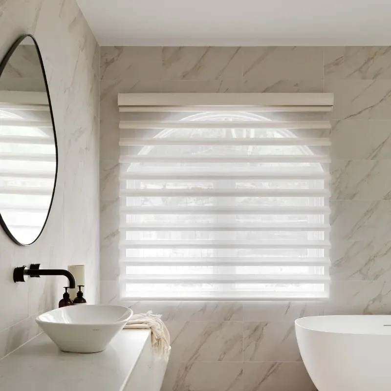 Bathroom with a white sink, black faucet, round mirror, and blinds.