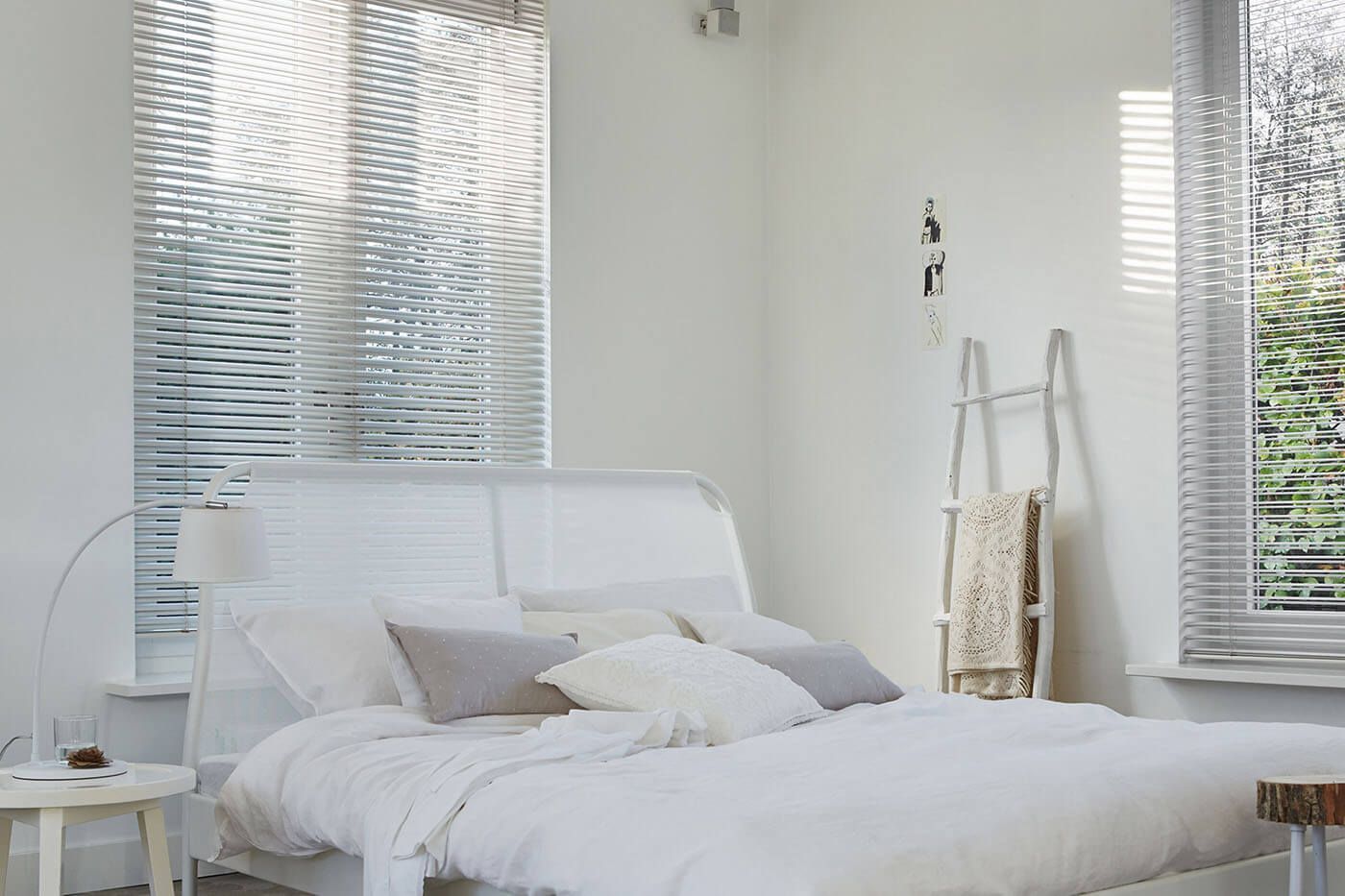 White bedroom with bed, pillows, windows with blinds, and ladder with blanket.