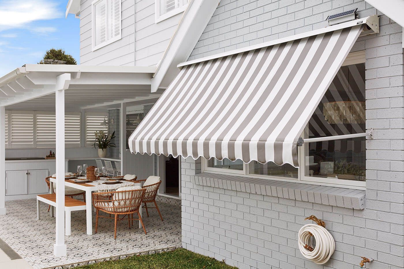 Grey and white striped awning over a window of a grey brick house. A table and chairs are on the patio.