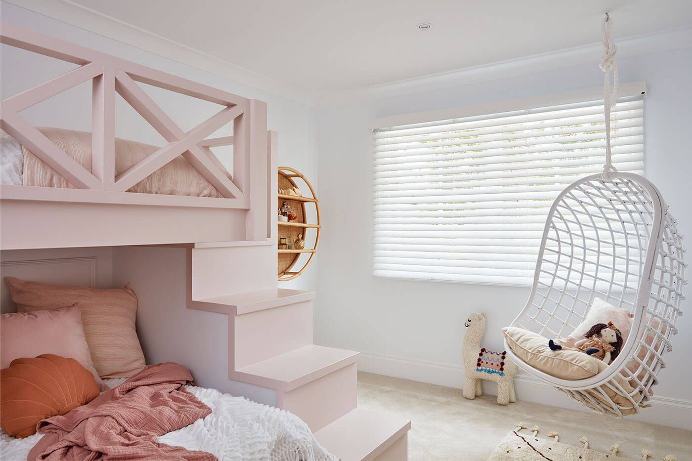 A pink kids' room with a loft bed, hanging chair, and round shelving unit by a window.