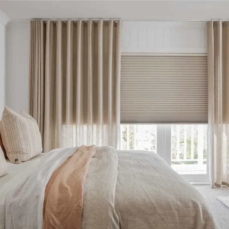 Bedroom with bed and window covered by blinds and beige curtains.
