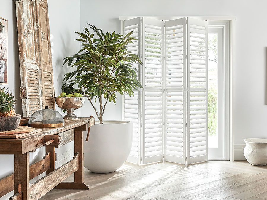 A bright room with a large white potted plant, folding shutters, and a rustic wooden table.