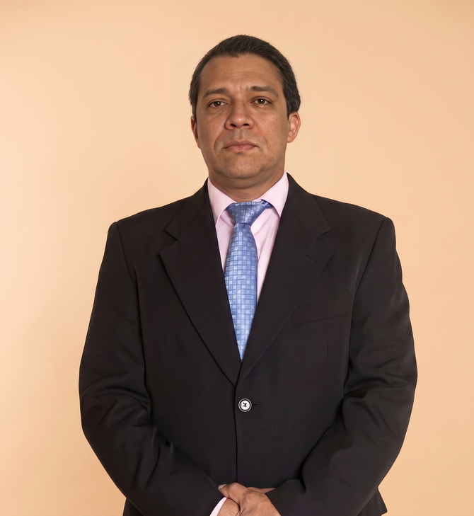 Man in a dark suit and blue tie standing against a plain beige background.