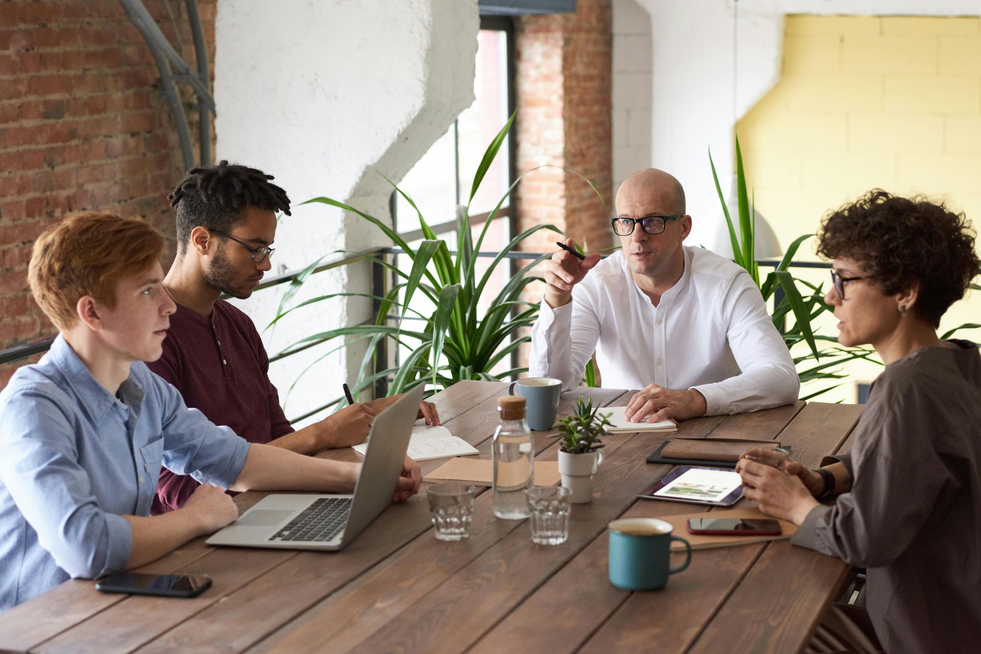 Four people meeting around a wooden table with laptops in a bright office