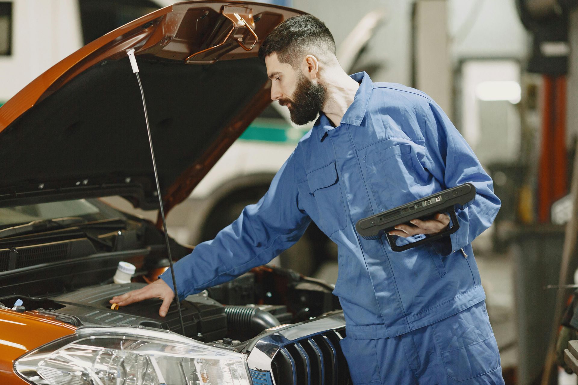 Mechanic in blue coveralls inspecting an orange car with the hood open in a garage
