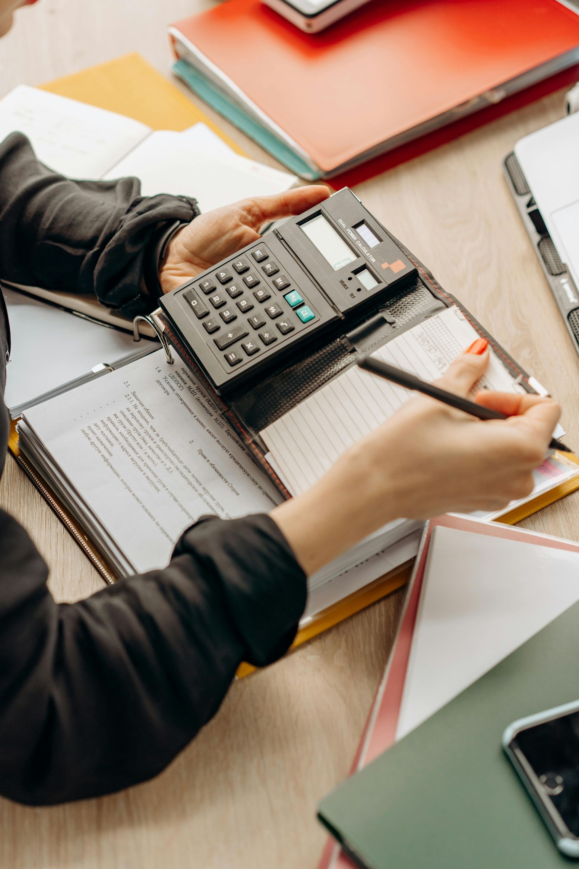 Person using a calculator beside paperwork at a desk with a red notebook and laptop, writing notes