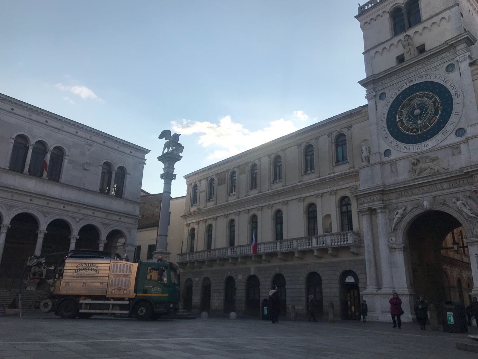Una piazza pubblica in Italia, con una torre dell'orologio, edifici, una colonna e un camion. Giornata di sole.