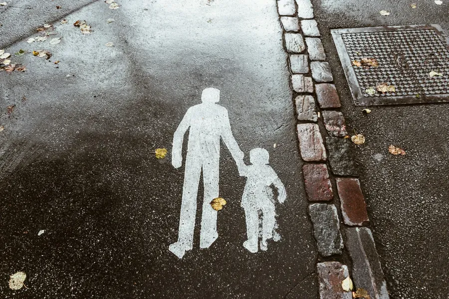 White pedestrian crossing symbol on wet pavement: adult holding a child’s hand.