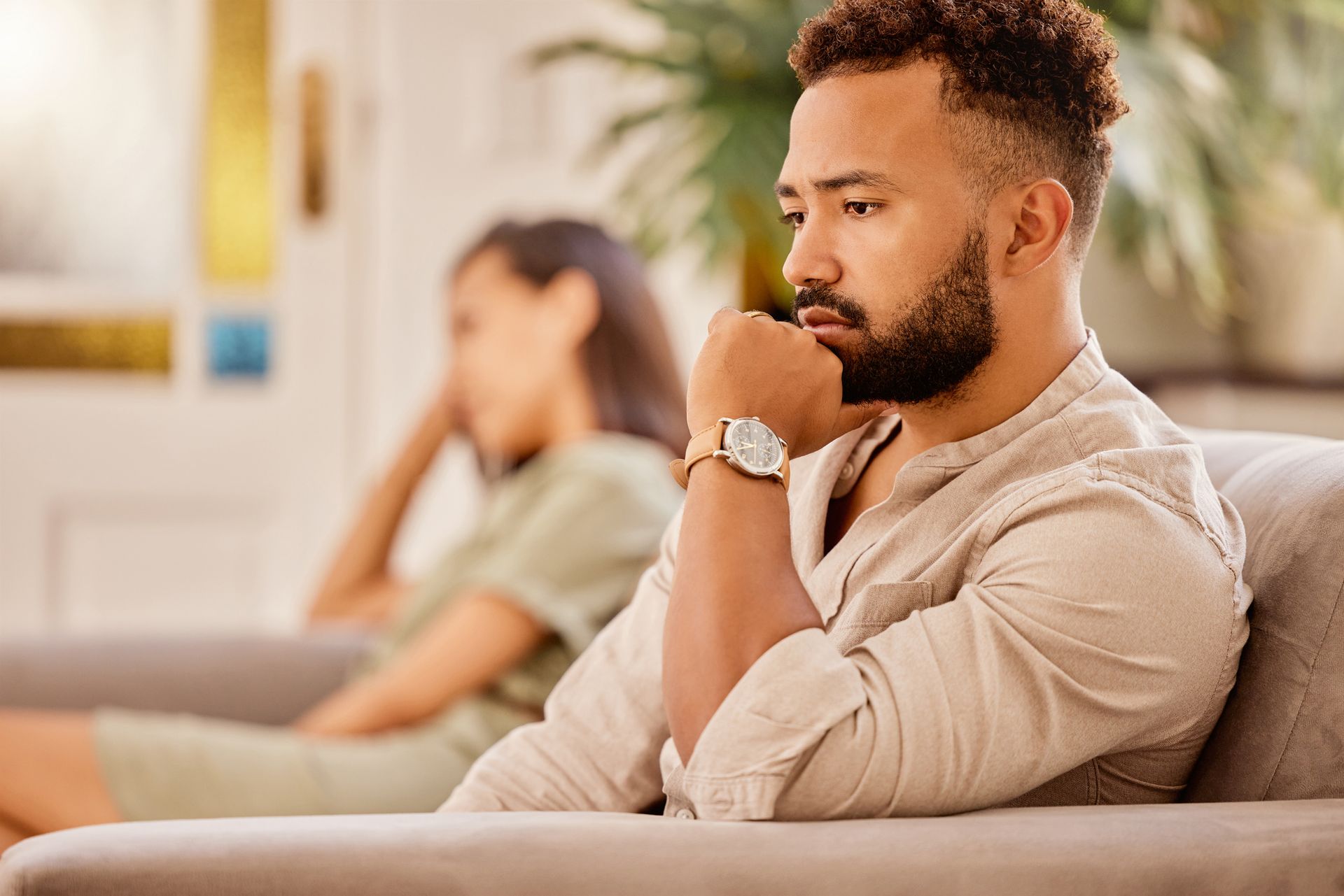 a man sitting on a couch, with a woman sitting back in the distance
