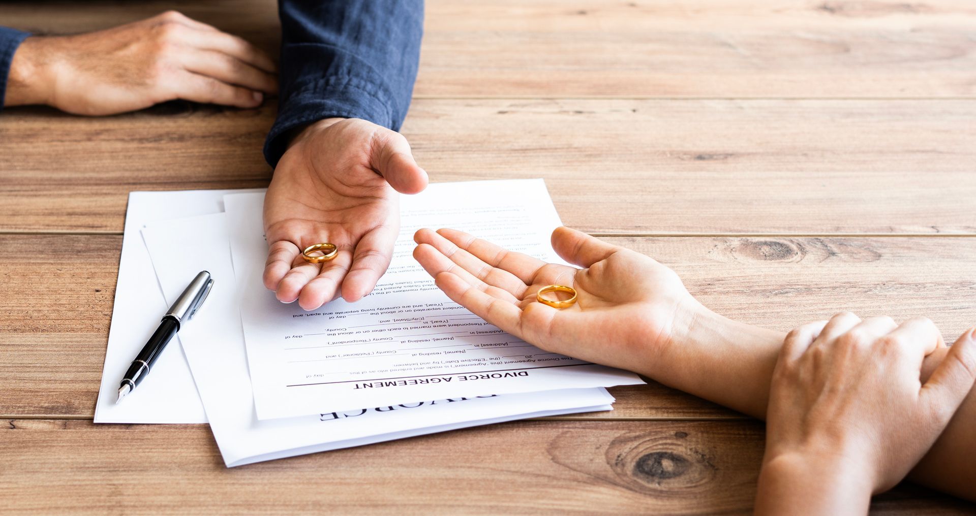 Hands exchanging wedding bands over paperwork on a wooden table with a pen nearby
