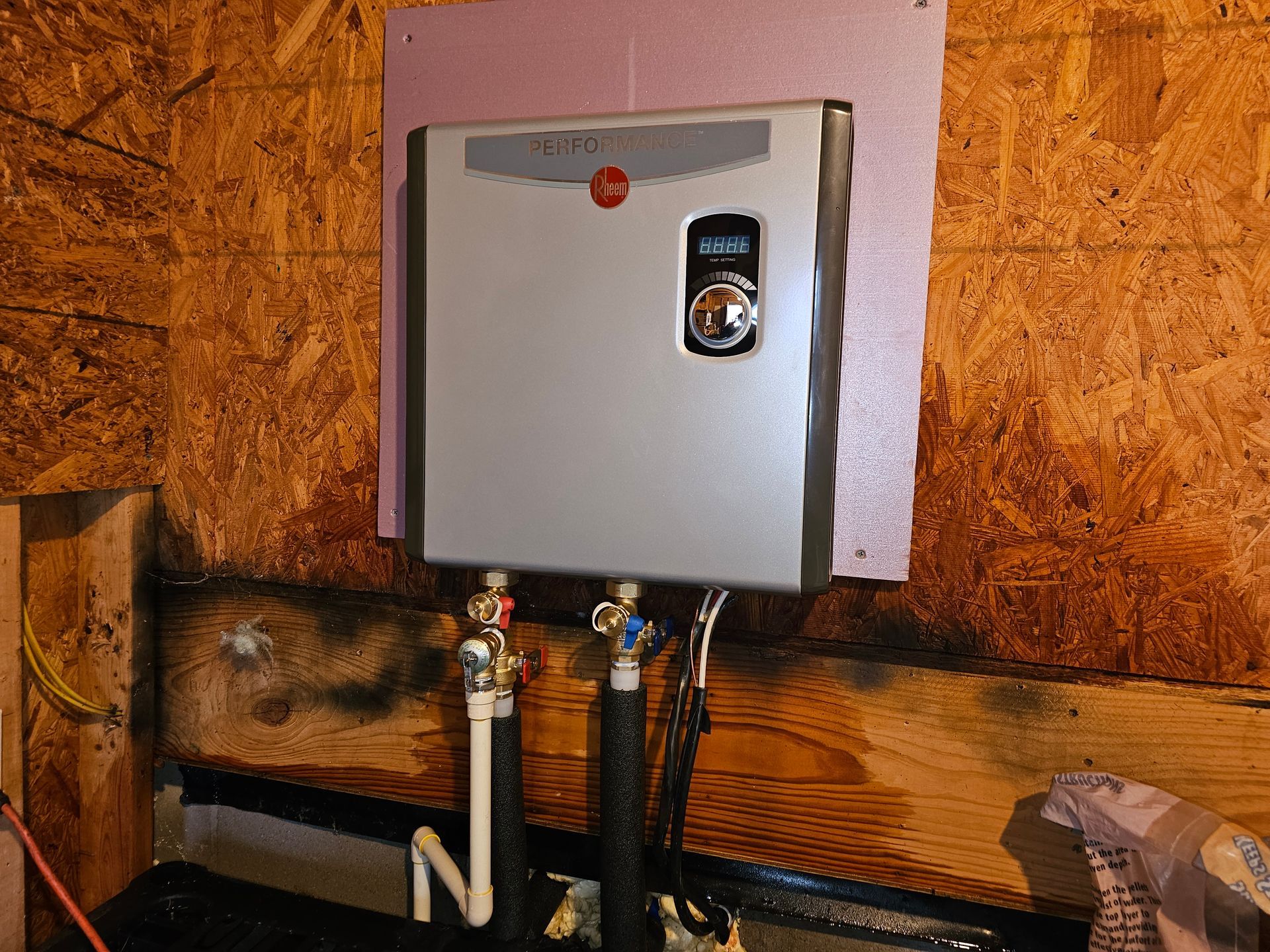 Tankless water heater mounted on a wooden wall, with pink insulation and water pipes.
