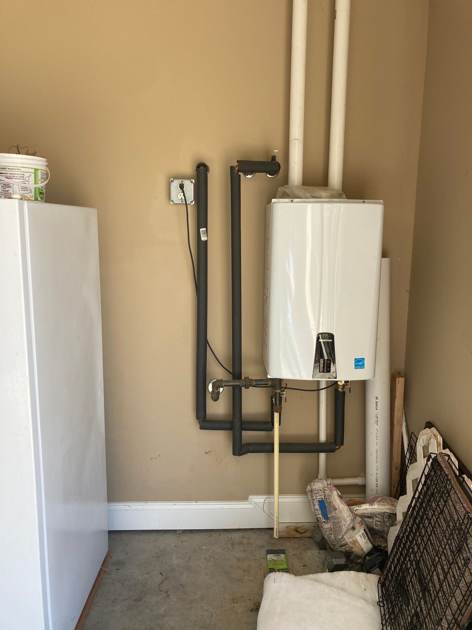 A tankless water heater installed on a beige wall in a garage, with connecting pipes and vent.