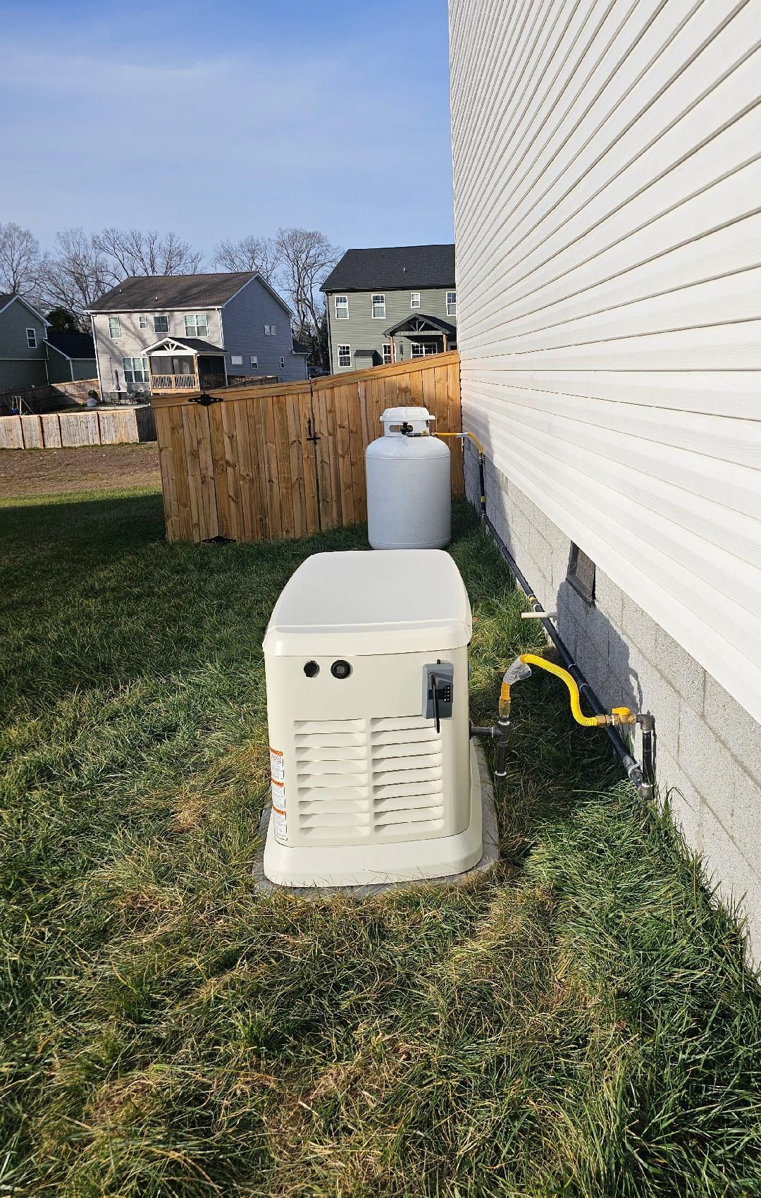 A standby generator and propane tank next to a house with wooden fence.