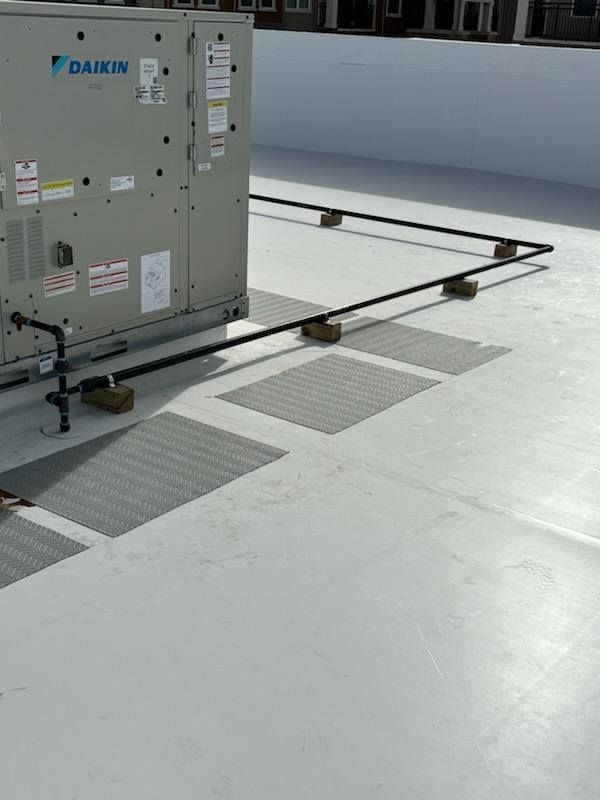 A rooftop HVAC unit with black piping elevated on wood blocks, with a white roof and metal ventilation grates.