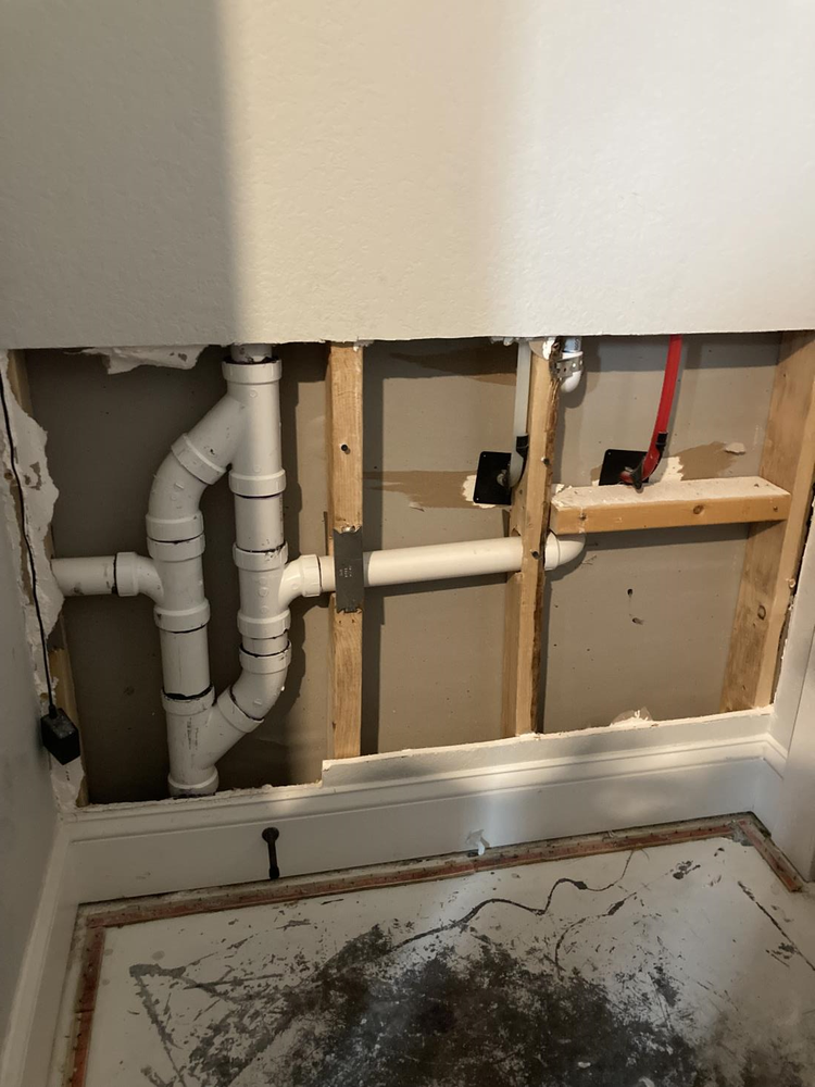 White plumbing pipes in an open wall cavity, including a p-trap and drain lines, next to wood framing.