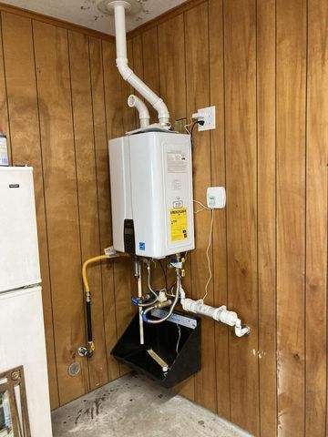 Wall-mounted white tankless water heater with vent pipe, drain pan, and gas line on wood paneling.