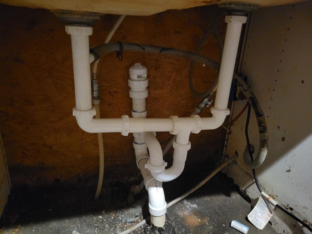 White PVC plumbing pipes under a sink. Horizontal and vertical pipes connect, with a central trap.