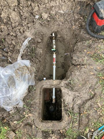 Open underground box with water pipes and valve in a muddy yard.